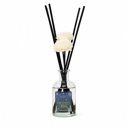 A reed diffuser bottle with a clear glass body, black reeds, and two white artificial flowers, with a label reading 'FROSTED ORRIS'.