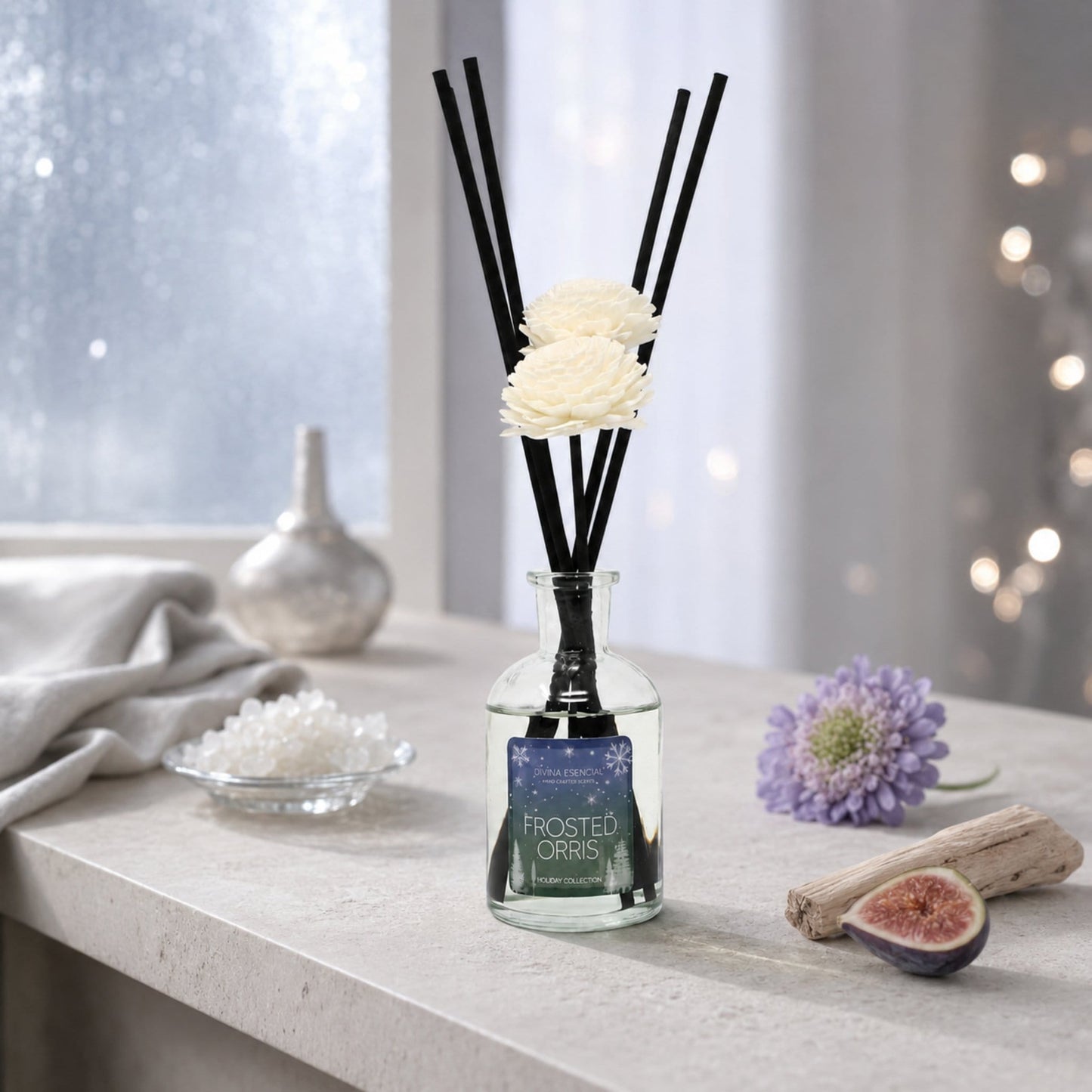 A frosted glass reed diffuser bottle with black reeds and cream flower pieces inside, labeled 'Frosted Orris,' set on a light stone surface near a window with decorative props including a purple flower, fig half, driftwood, and a small dish of crystals.