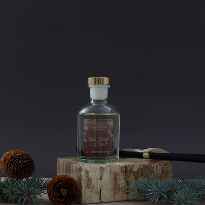 Bottle of Balsam Clove on a wooden block with pine cones and branches against a dark background