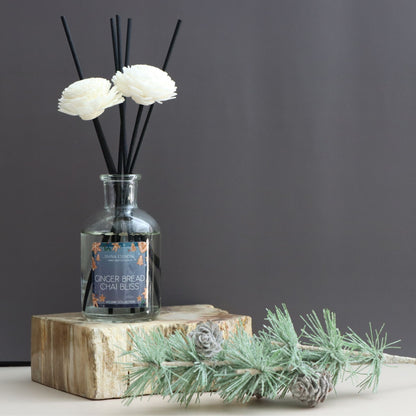 Diffuser bottle with white flowers and black sticks on a wooden block against a gray background