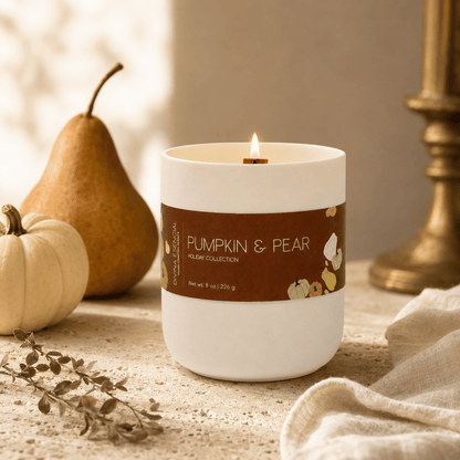 A white jar candle with a lit wick sits on a tabletop in a styled autumn scene. The candle has a brown label reading 'PUMPKIN & PEAR' and 'Holiday Collection.