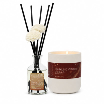 A reed diffuser bottle with black reeds and two white flowers next to a lit white candle jar labeled Sparkling Winter Vanilla on a white background.